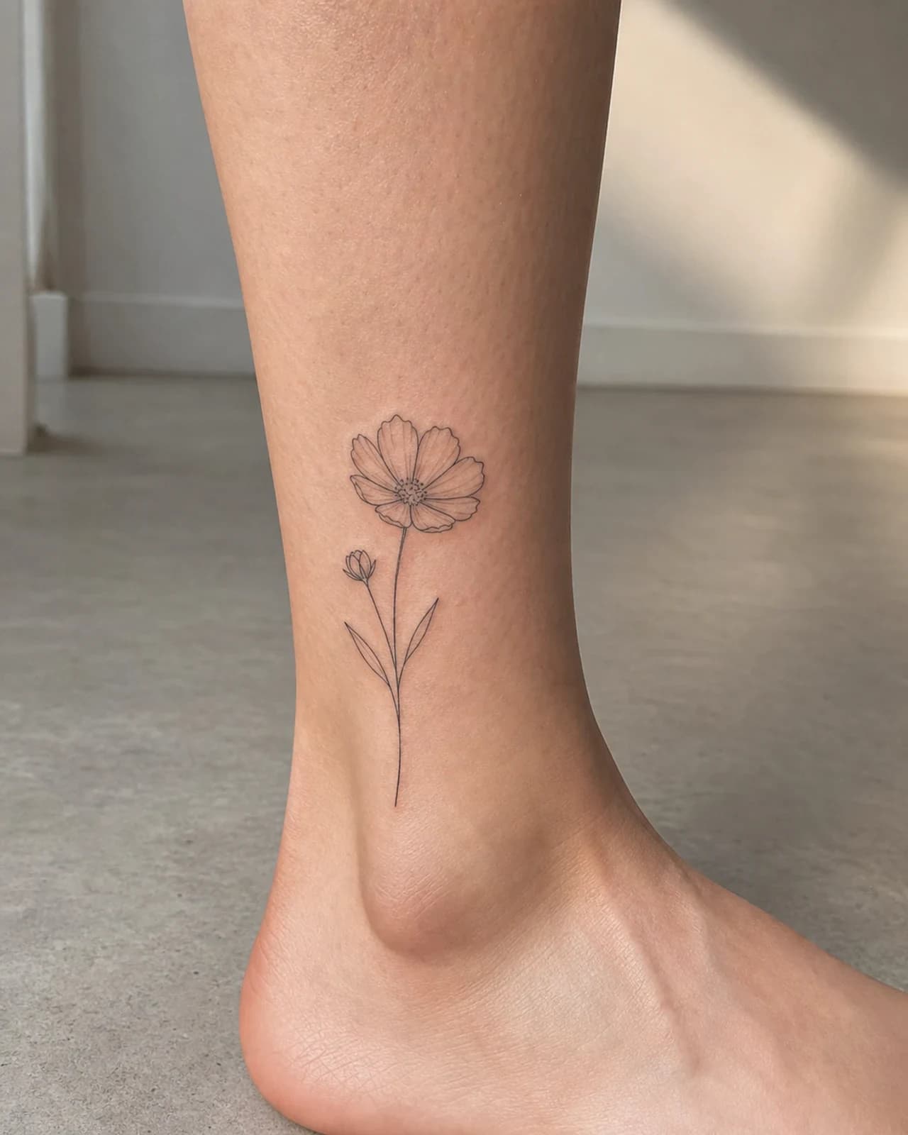 Fine line cosmos flower sprig tattoo positioned above an adult outer ankle bone