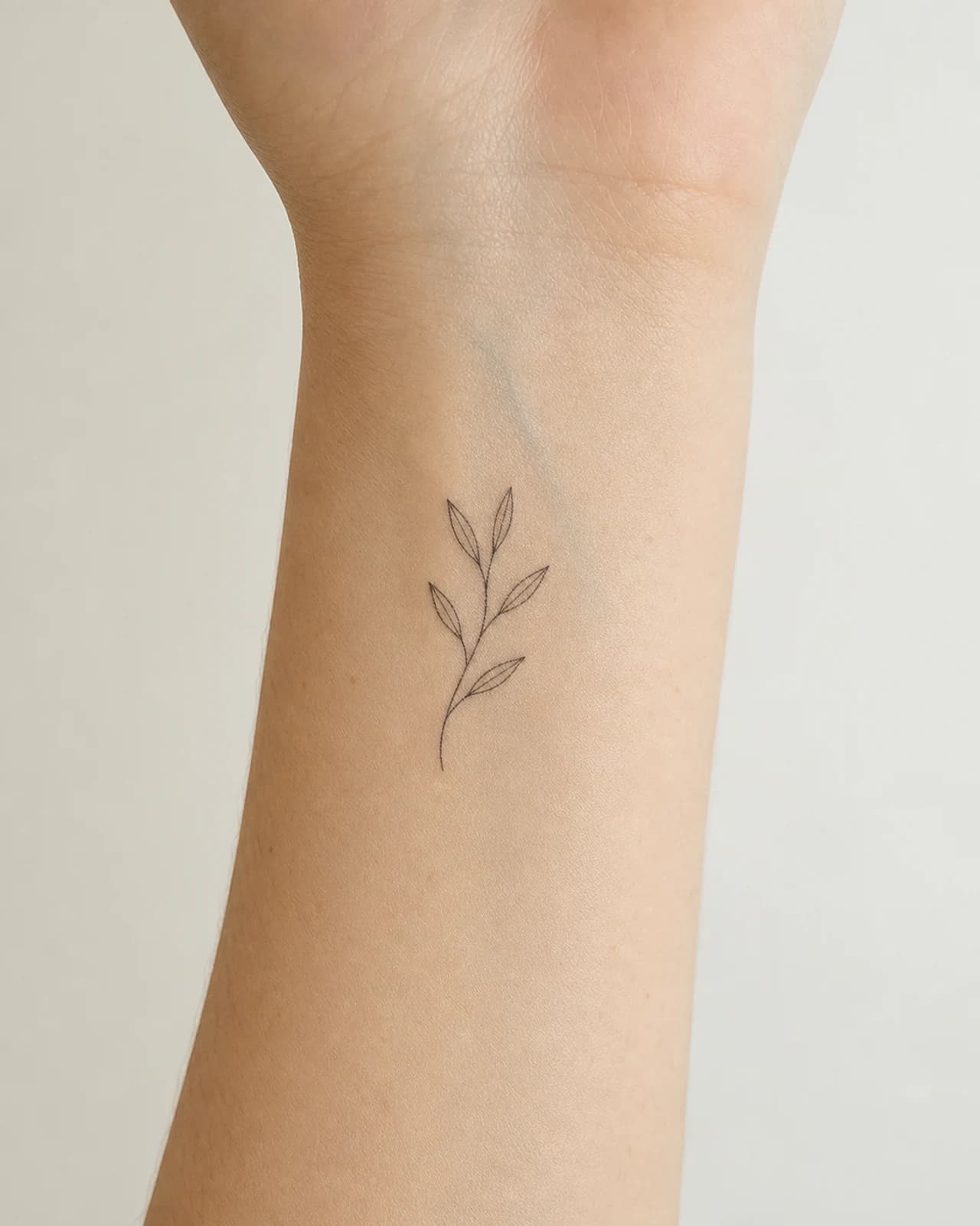 Small black fine line olive sprig tattoo placed above an adult inner wrist crease