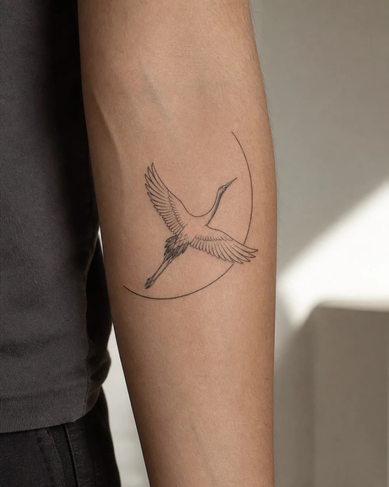 Minimal fine line crane with an orbit arc tattooed vertically on an adult inner forearm